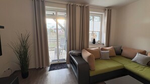 A living room with a green sofa, beige curtains, and a potted plant. - Haus am Röblinsee - Stork vacation apartment in Fürstenberg / Havel (Fürstenberg/Havel)