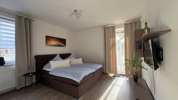 A bedroom with a bed, a TV mounted on the wall, a window with curtains, and a plant. - Haus am Röblinsee - Stork vacation apartment in Fürstenberg / Havel (Fürstenberg/Havel)