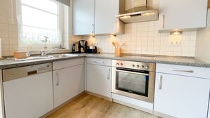 A kitchen with white cabinets, a built-in oven, a coffee maker, and a window with a view. - Tannengrund - Comfortable vacation apartment little time out with terrace and garden (Loddin)