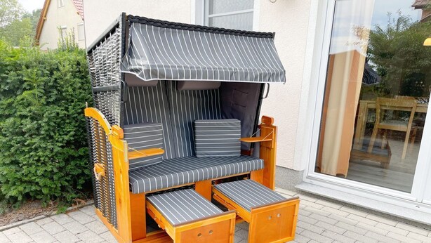 A wooden outdoor seating area with a striped awning, a small table, and a glass door leading to a patio. - Tannengrund - Comfortable vacation apartment little time out with terrace and garden (Loddin)