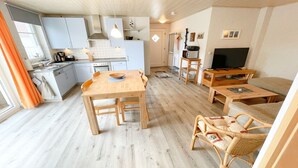 A modern kitchen with white cabinets, a dining table with chairs, a wicker sofa, a TV, and a wooden floor. - Tannengrund - Comfortable vacation apartment little time out with terrace and garden (Loddin)