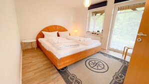 A bedroom with a wooden bed, white bedding, a small bedside table, a hanging light, and a view through a window. - Tannengrund - Comfortable vacation apartment little time out with terrace and garden (Loddin)