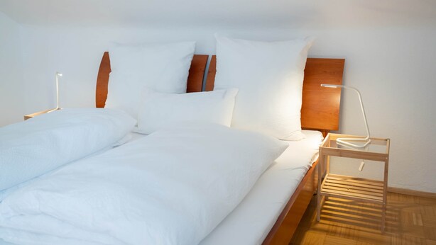 A neatly made bed with white linens and two pillows, a bedside table with a lamp, and a wooden nightstand. - Guesthouse Elena Apartment 13 (Kassel)