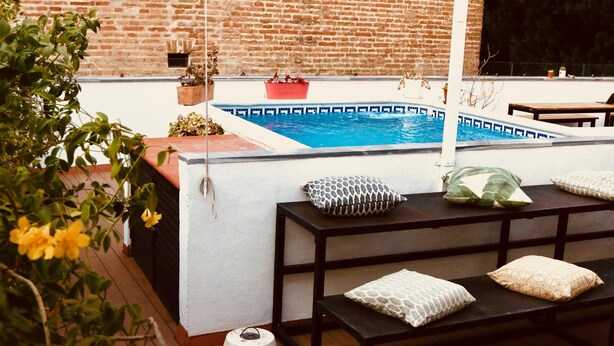 Zona de piscina con pared de ladrillo, terraza de madera y área de estar con cojines.