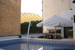 A pool area with a tiled border, a brick wall, and a building with a patterned roof in the background. - Joy Setas Coworking (Seville)