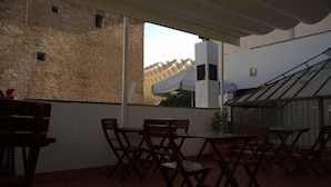 An outdoor patio with wooden tables and chairs, a brick wall, and a view of a modern building with a sloped roof. - Joy Setas Coworking (Seville)
