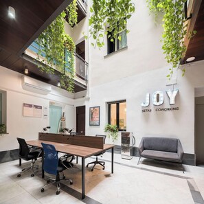 A modern office space with a wooden table, office chairs, a grey sofa, and hanging plants. - Joy Setas Coworking (Seville)