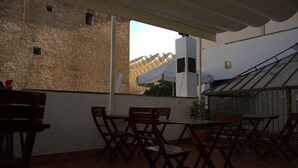 Un patio al aire libre con mesas y sillas de madera, un muro de ladrillo y vistas a un edificio moderno con tejado inclinado.