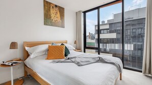 A modern hotel room with a large bed, a side table with a lamp, a painting on the wall, and a view of the city through large windows. - Luxury Living in Heart of South Yarra near Jam Factory, Prahran Market Chapel St (South Yarra)