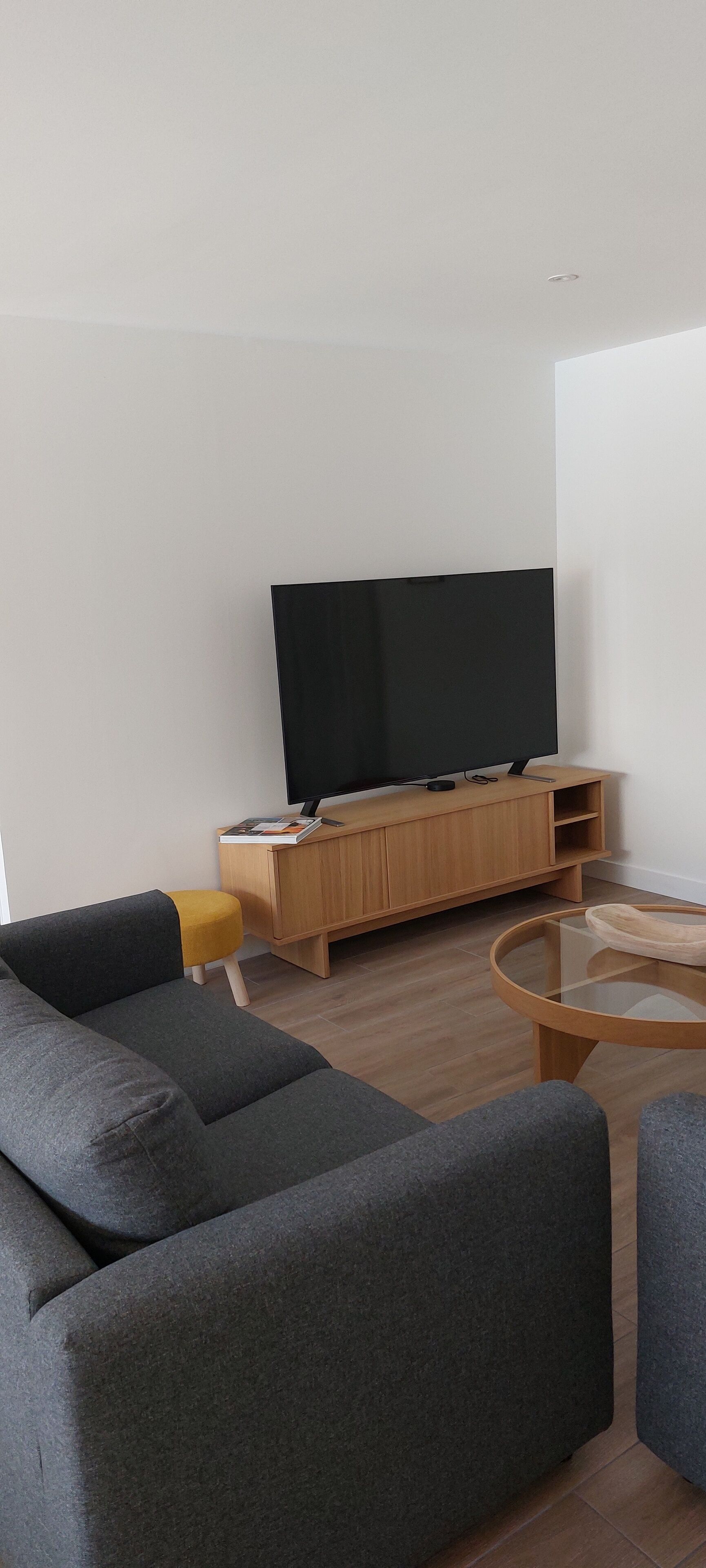 A modern living room with a large flat-screen TV, a wooden entertainment unit, a round glass coffee table, and two grey sofas.