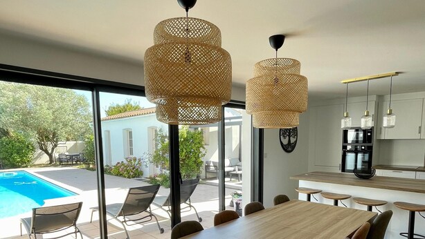 Eine moderne Küche mit einem großen Esstisch, zwei hängenden Rattan-Kronleuchtern und Blick auf einen Swimmingpool.