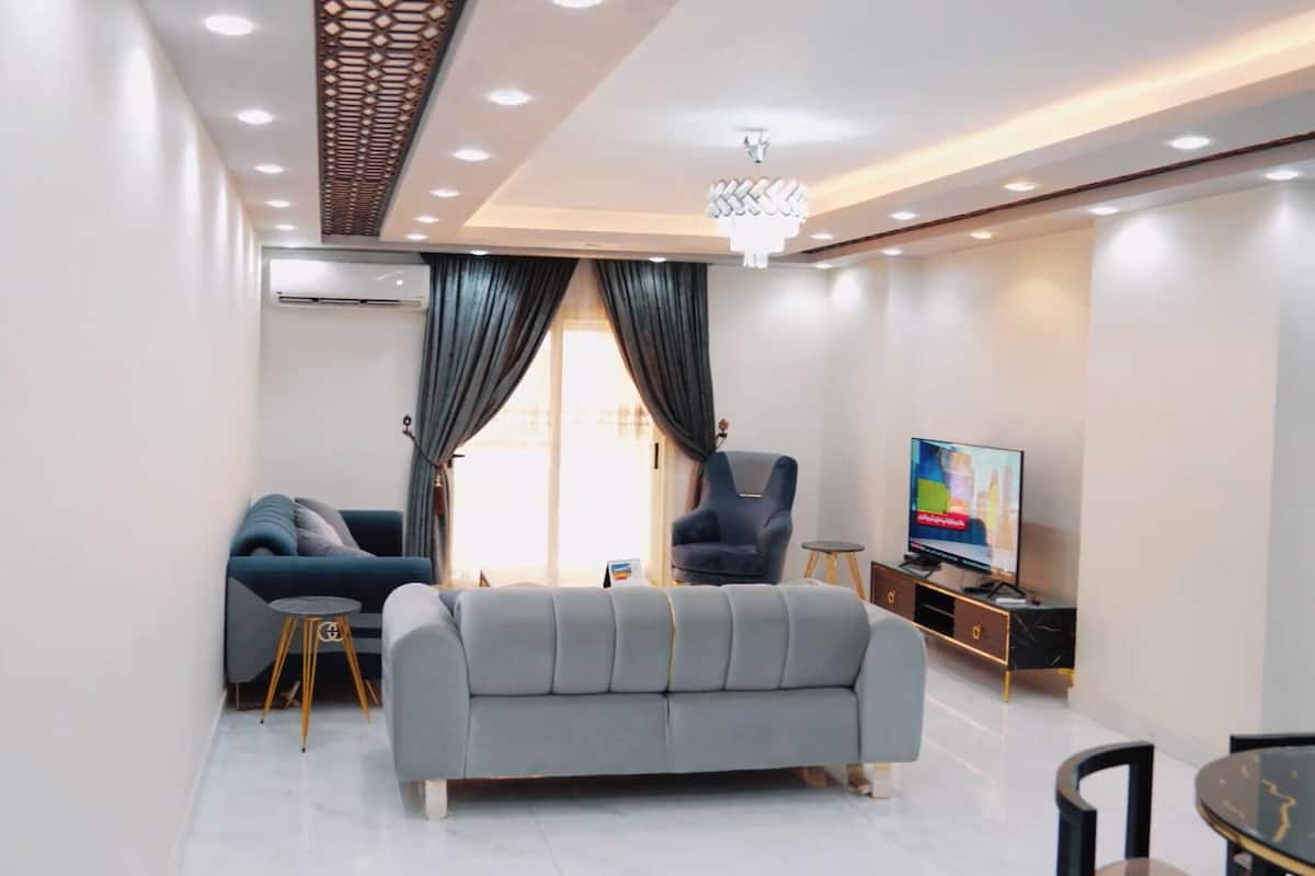 A modern living room with a grey sofa, a blue armchair, a small round table, and a television on a wooden stand.