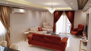 Ein modernes Wohnzimmer mit einem roten Sofa, beigefarbenen Sesseln, einem Glastisch und einer Deckenleuchte.