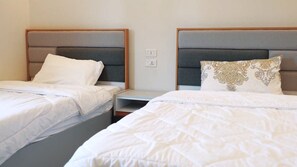 Ein Hotelzimmer mit zwei Betten, jeweils mit Kopfteil und Kissen. Dazwischen befindet sich ein Nachttisch und an der Wand eine Steckdose.