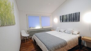 A bedroom with a bed, a desk, a chair, a radiator, and two windows.