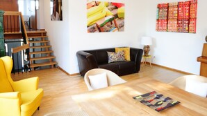A living room with a wooden staircase, a black sofa, a yellow armchair, a dining table, and a colorful artwork on the wall.