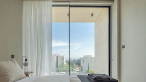 A bedroom with a bed, a bedside table, a lamp, a window with sheer curtains, and a view of buildings and greenery outside.