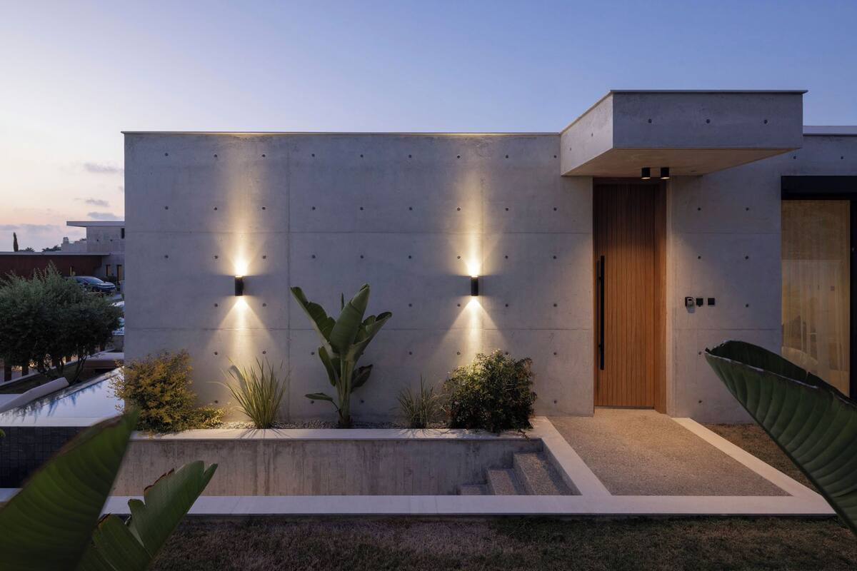 A modern building with a concrete facade, two illuminated wall lights, a wooden door, and a small garden area with plants.