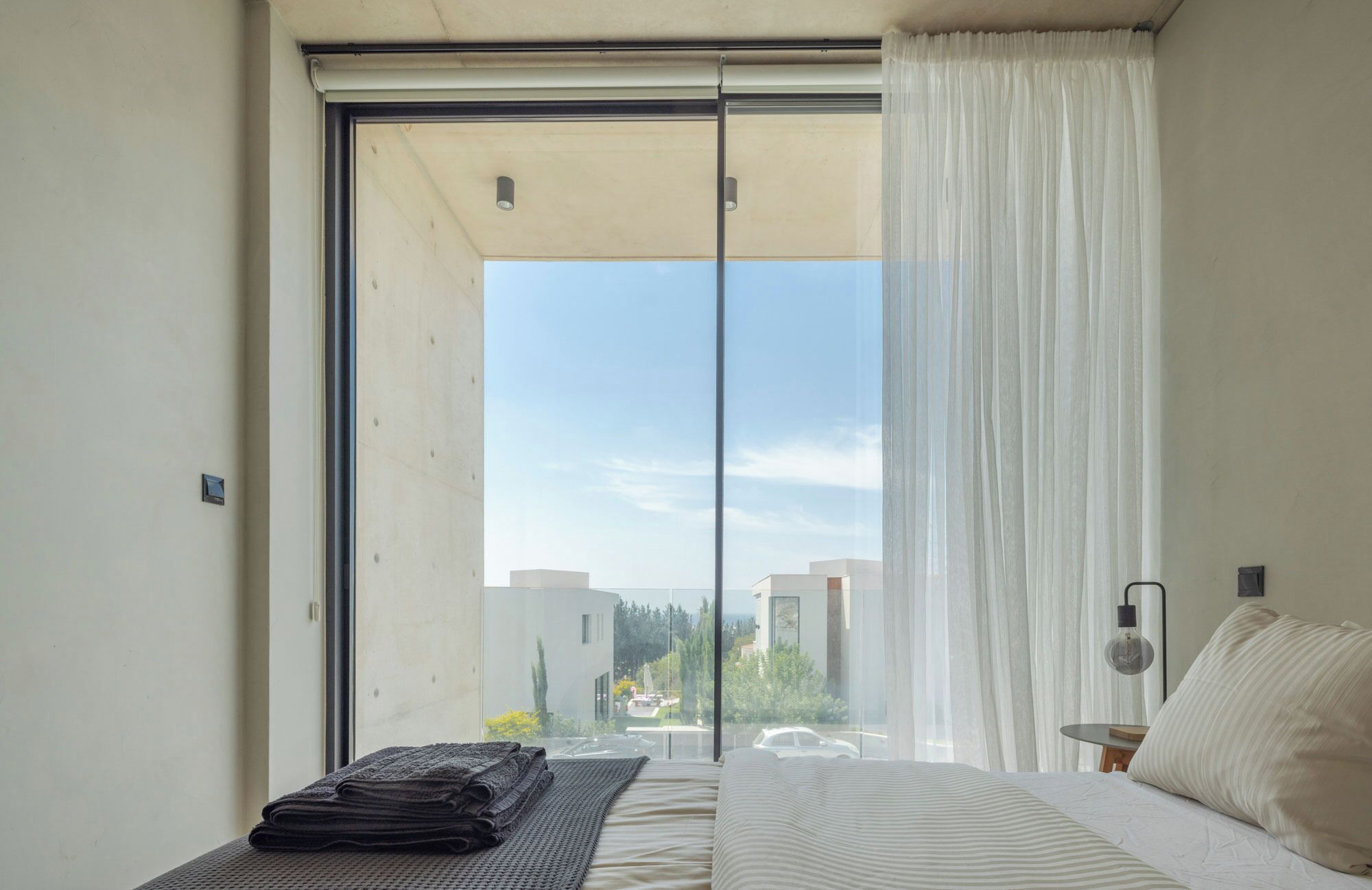 A bedroom with a large sliding glass door leading to a balcony, a bed with a white and beige striped bedspread, a small bedside table with a lamp, and a stack of folded towels on a mat.