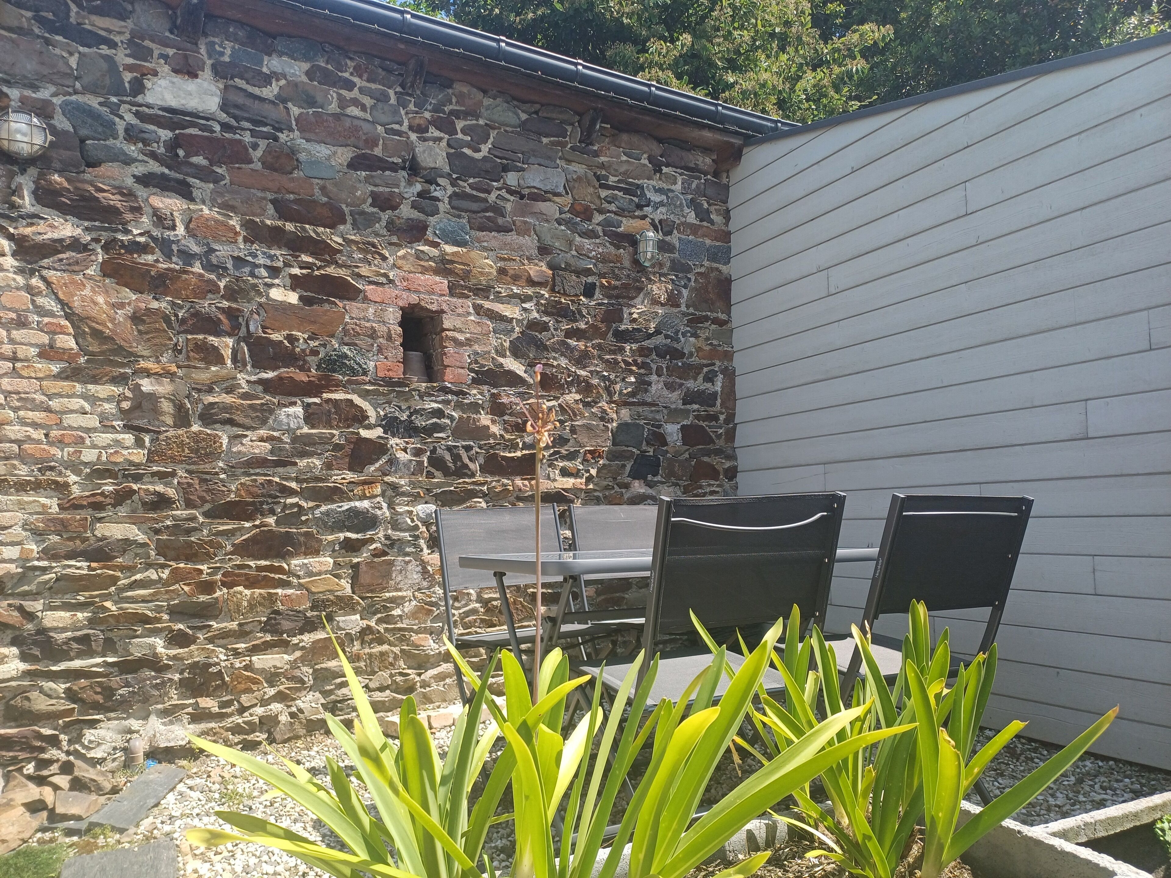 Un mur de pierre avec une petite fenêtre, une table de patio avec des chaises et des plantes vertes devant.