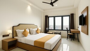 A hotel room with a large bed, a desk, a chair, a TV, and a view of the city through the window. - TGR Hotel (Mundra)