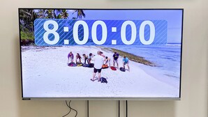 A television displaying a beach scene with a timer at 8:00:00, a Scrabble box, two game controllers, a green toy, and a small plant on a wooden table.