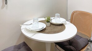 A round white table with a woven placemat, two clear glass tumblers, a small potted plant, and a metal spoon. A grey cushioned bench is in front of the table. A wicker chair with a black cushion is to the right. A white wall-mounted heater is visible on the left.