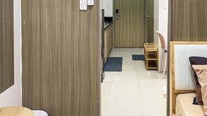 A hotel room with a bed, a small desk, a chair, a trash bin, and a door leading to a bathroom and kitchen area.
