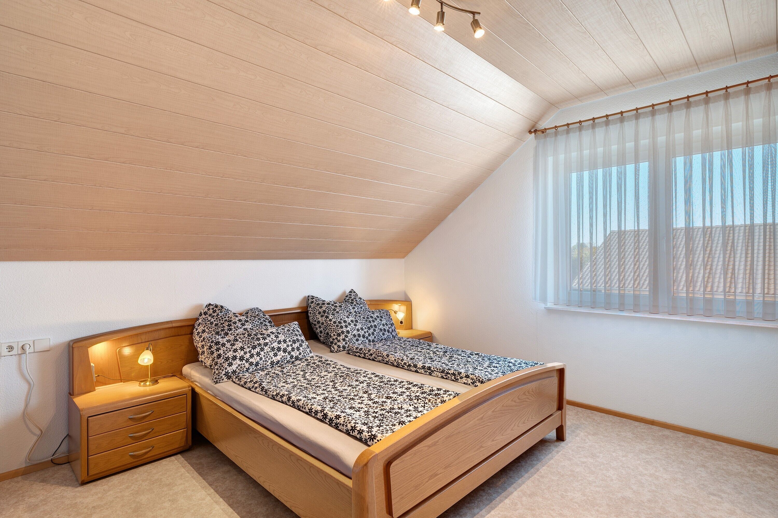 A bedroom with a sloped ceiling, a wooden bed, bedside lamps, a nightstand, and a window with sheer curtains.