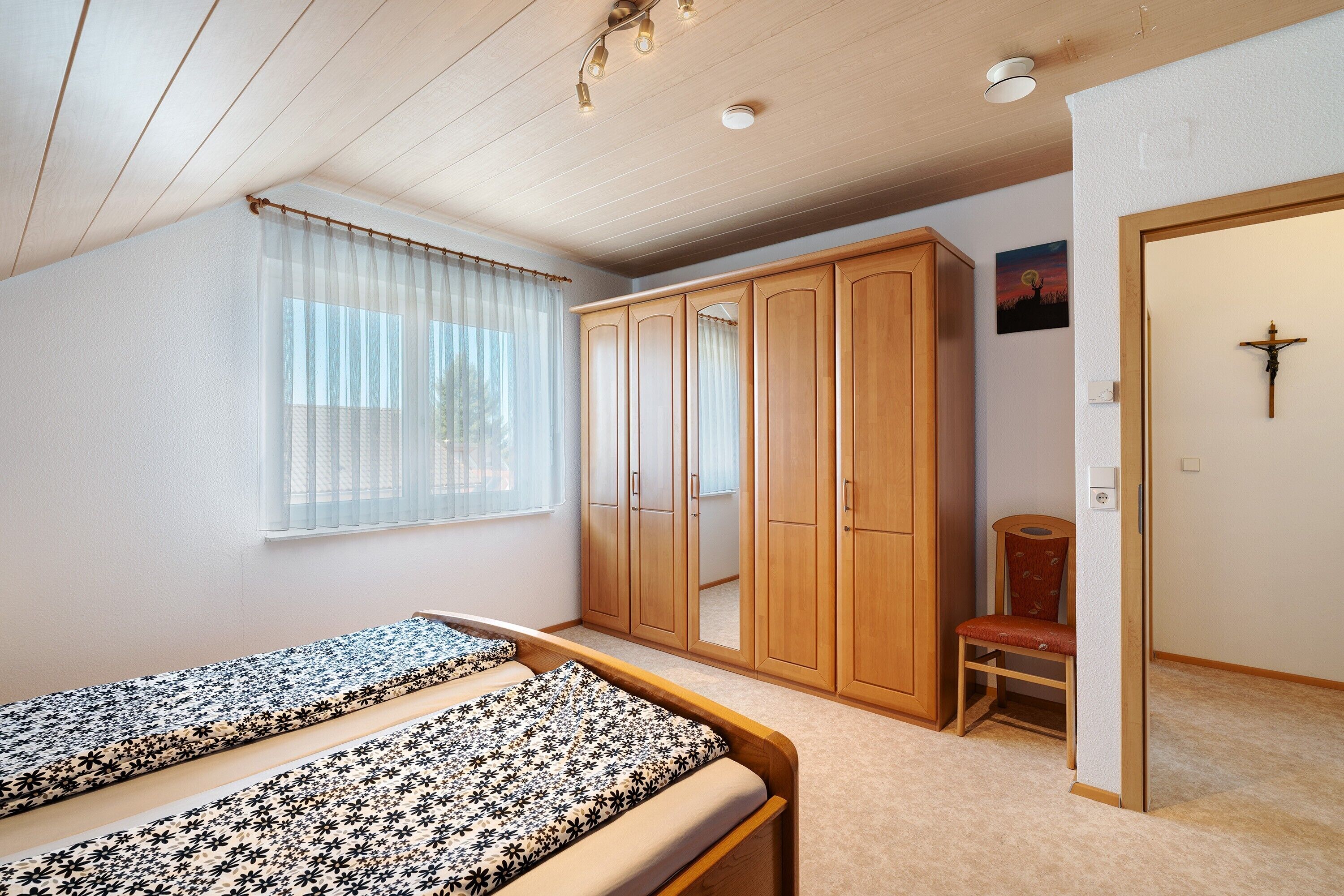 A bedroom with two beds, a wardrobe, a chair, and a cross on the wall.