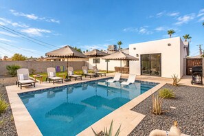 A swimming pool with lounge chairs and umbrellas, surrounded by a modern house with a patio.