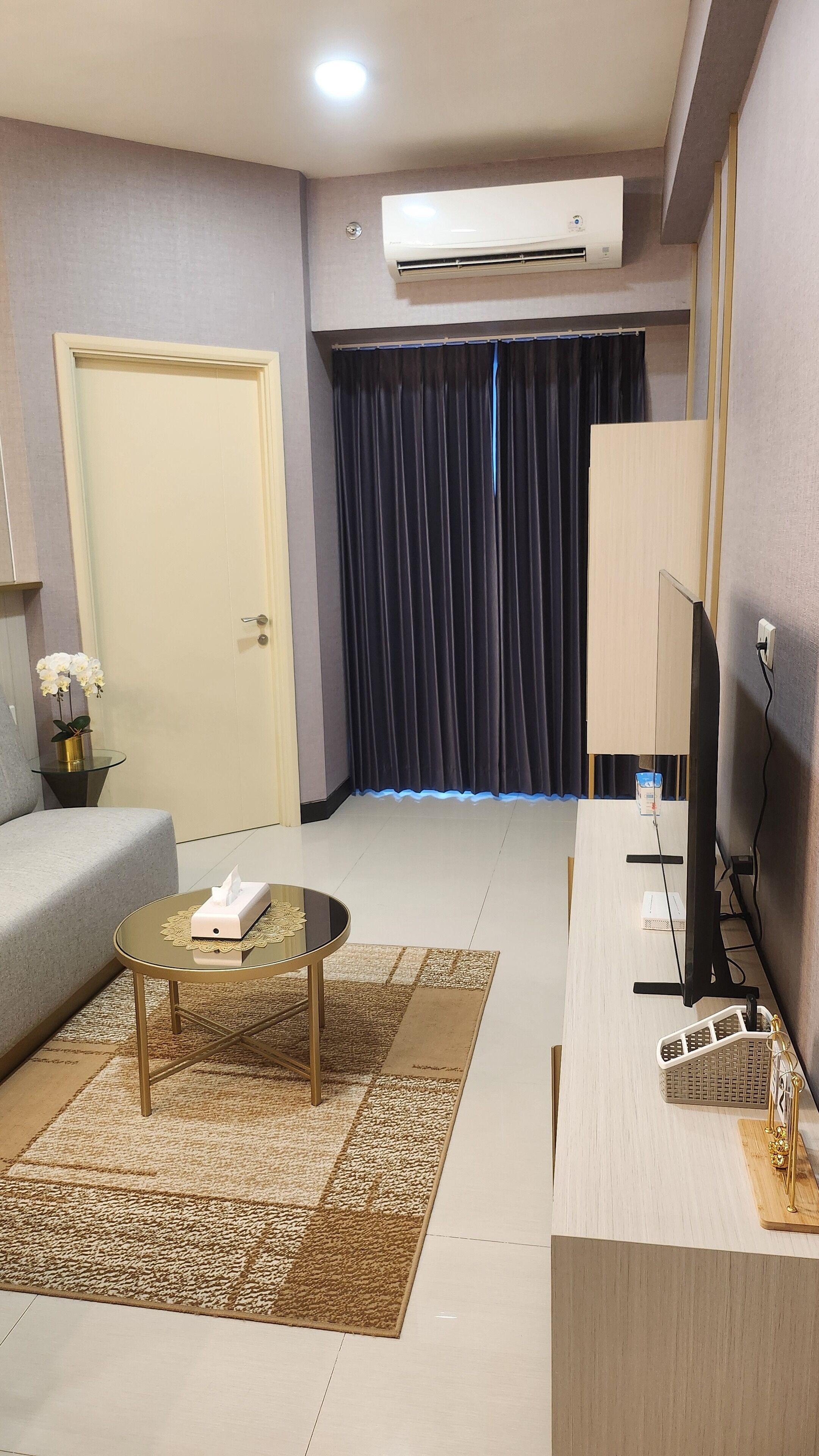 A hotel room with a sofa, a small round table with a tissue box, a television, a desk with a computer, and a large window with curtains.