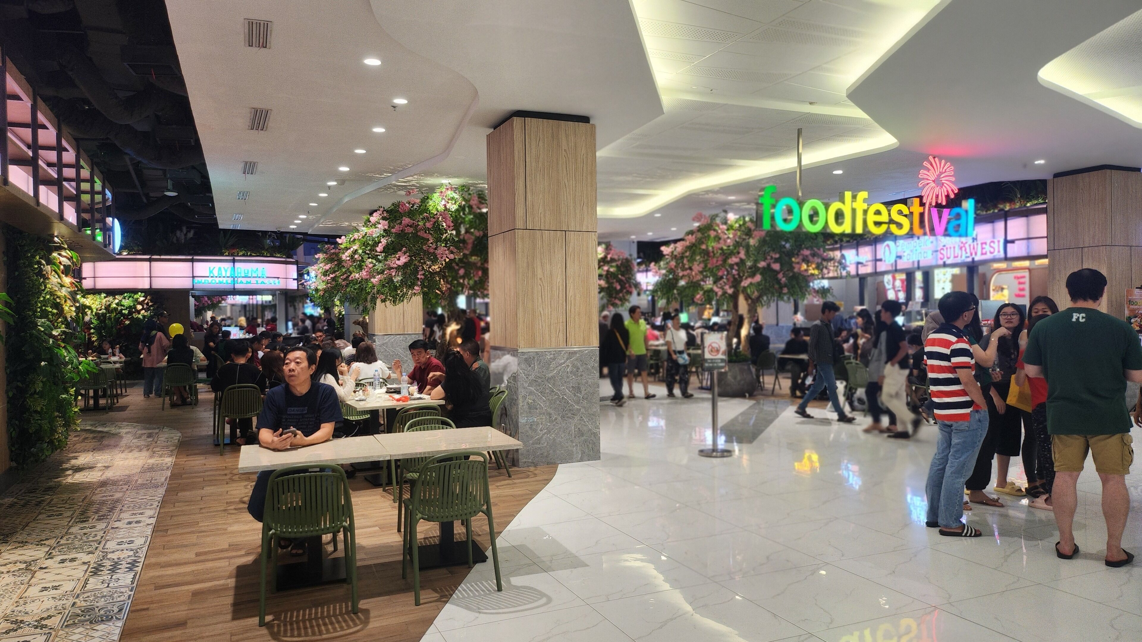 Un espace de restauration avec de nombreuses tables et chaises, entouré de différents stands de nourriture et d’une enseigne annonçant un « festival gastronomique ».