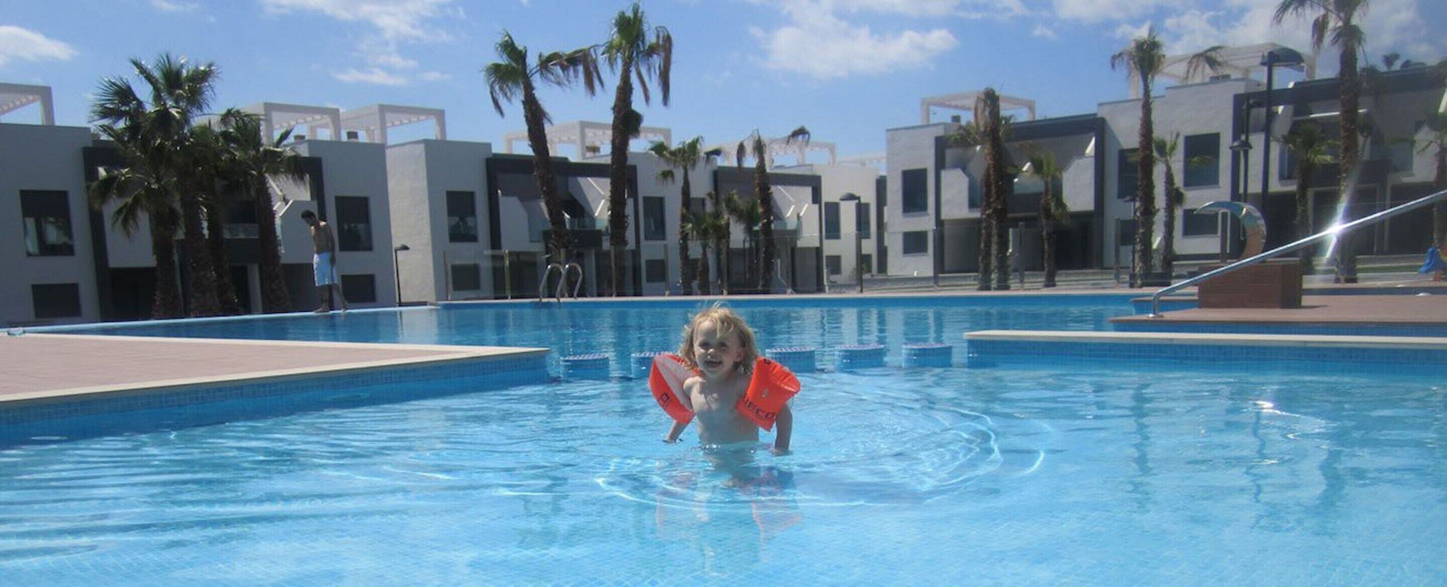 Et barn i en swimmingpool med moderne bygninger og palmer i baggrunden.