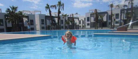 Niño en una piscina con edificios modernos y palmeras al fondo.