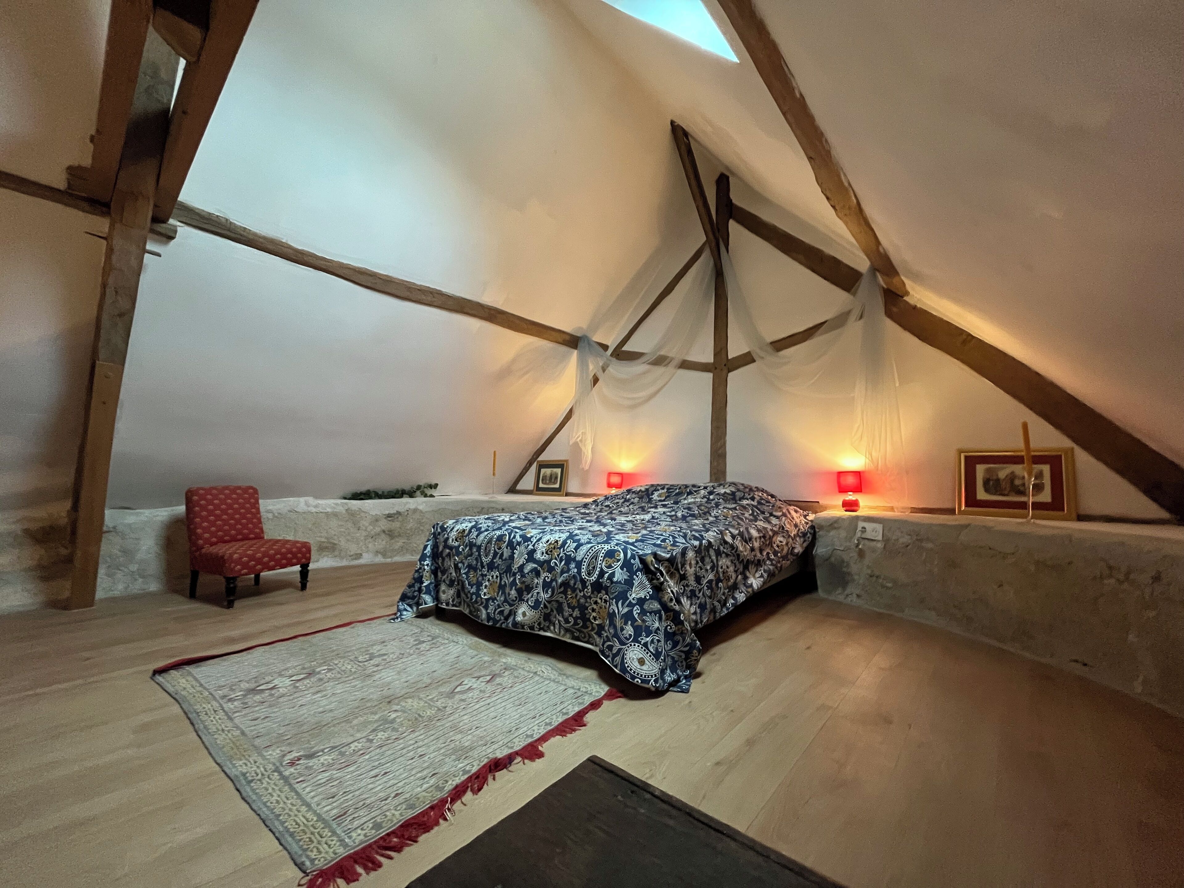 A cozy attic bedroom with a bed, a red chair, and two wall-mounted lamps.