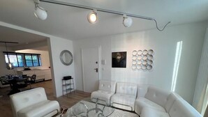 Un salon moderne avec une table basse en verre, des canapés blancs, un coin repas avec une table et des chaises, et une cuisine en arrière-plan.