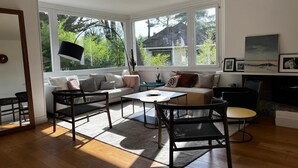 Un salon spacieux comprenant un canapé, des fauteuils, une table basse et une cheminée. De grandes fenêtres laissent entrer la lumière naturelle et offrent une vue sur l’extérieur.
