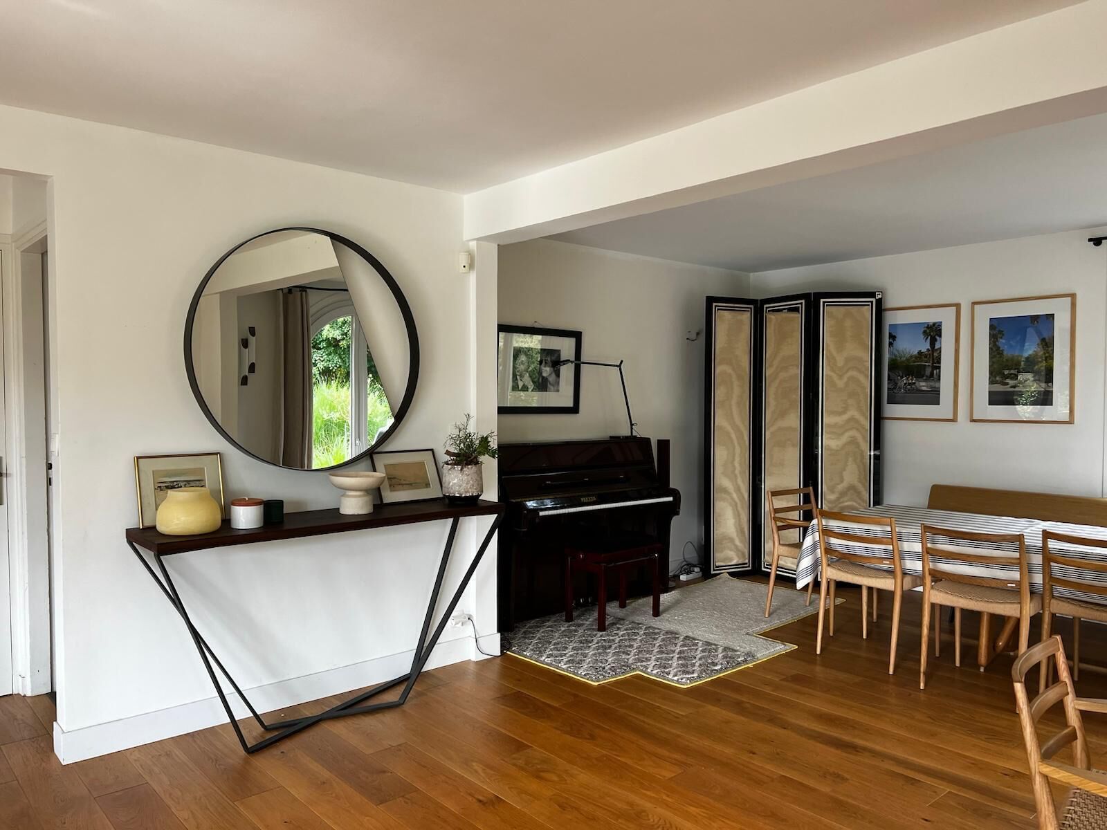 Un salon avec un piano, un grand miroir rond, une table à manger avec des chaises et des tableaux encadrés accrochés au mur.