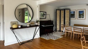 Un salon avec un piano, un grand miroir rond, une table à manger avec des chaises et des tableaux encadrés accrochés au mur.