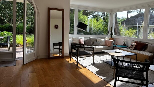 Une pièce lumineuse, avec un canapé, deux fauteuils, une table basse et un miroir. De grandes fenêtres offrent une vue sur la verdure environnante.