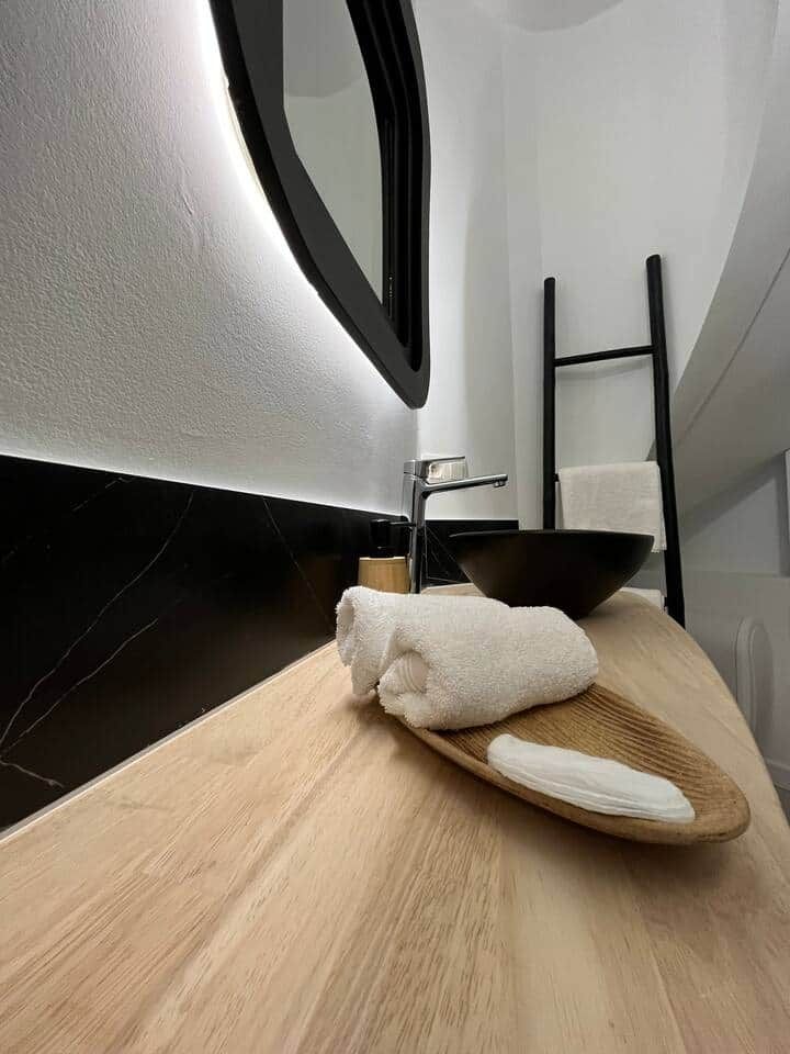 Une salle de bain avec un meuble-lavabo en bois, un lavabo noir, un miroir et une serviette posée sur un plateau en bois.
