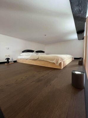 Een slaapkamer met een houten bed, een klein nachtkastje, een lamp en een wandcontactdoos.
