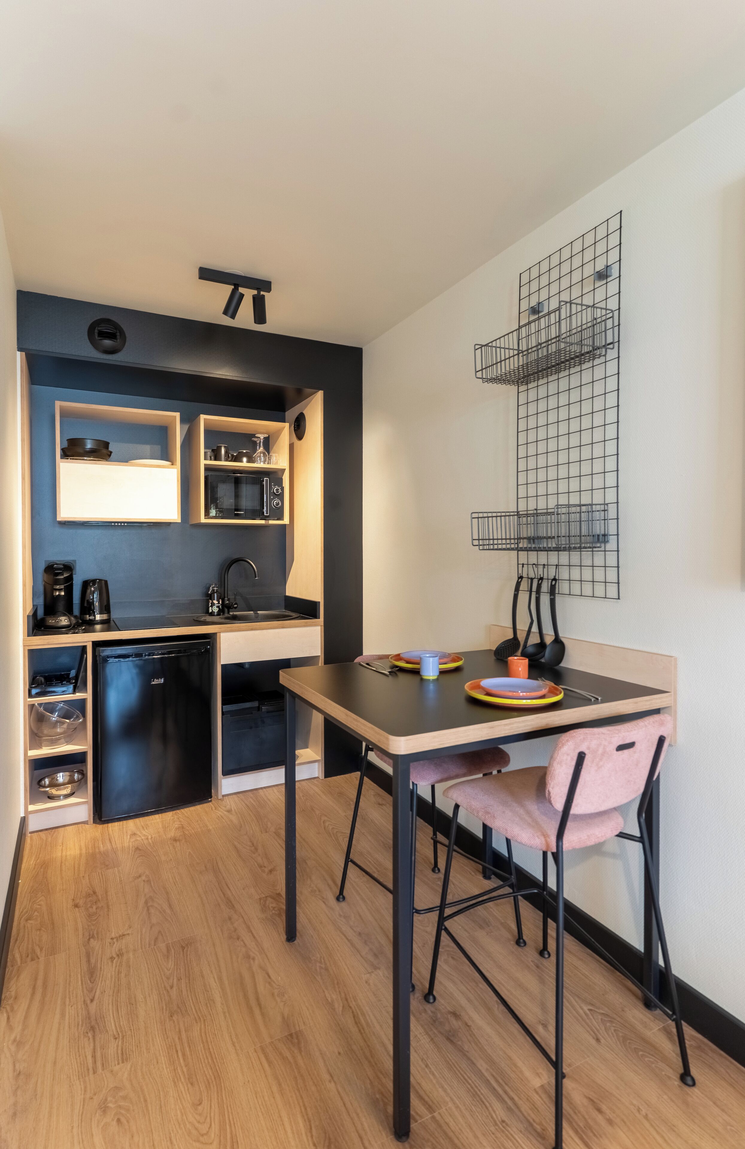 A compact kitchen with a microwave, coffee maker, and a small refrigerator. There is a dining table with a chair and a wall-mounted shelf above it.