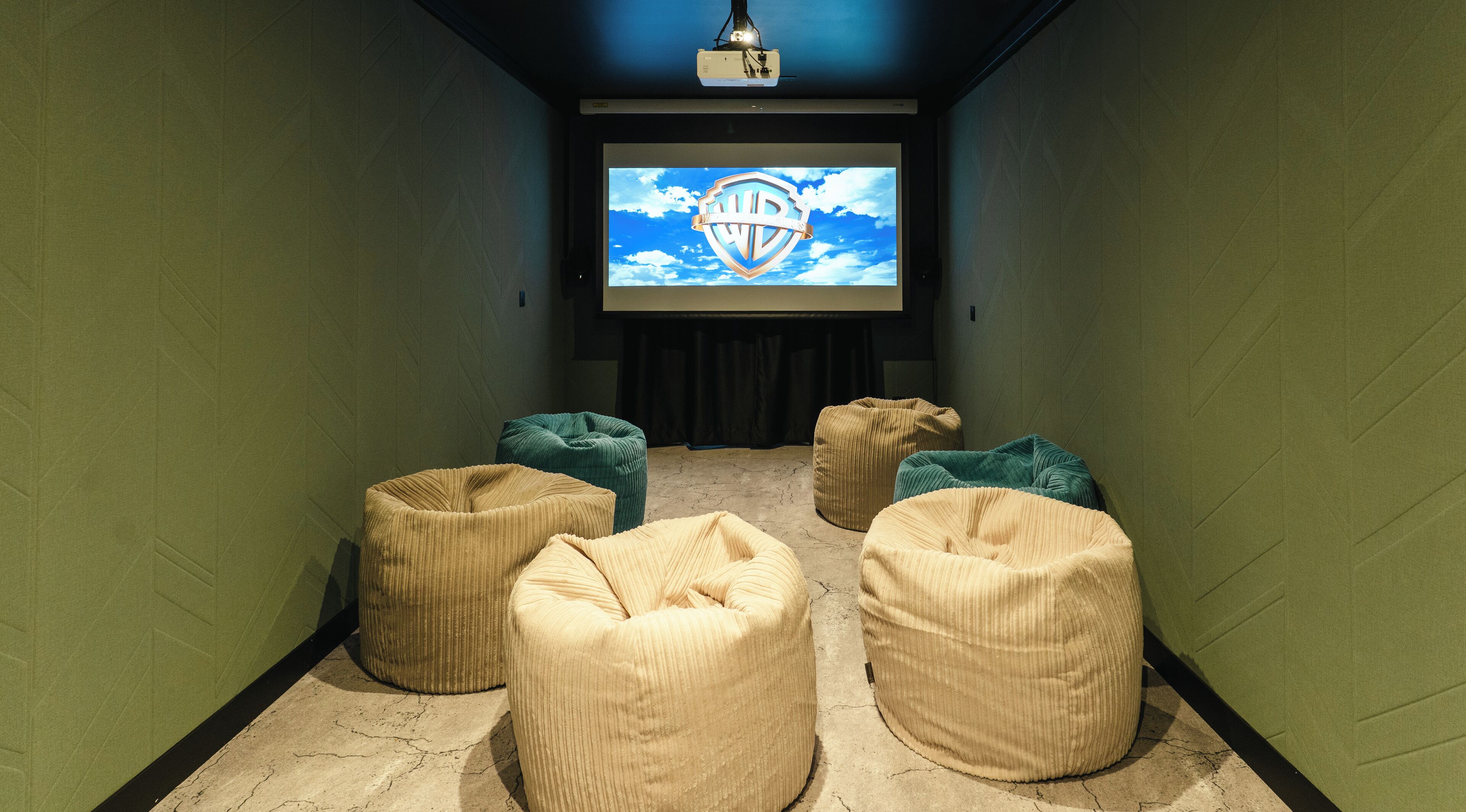 A cinema room with a large screen displaying the Warner Bros logo, surrounded by several beige bean bag chairs.