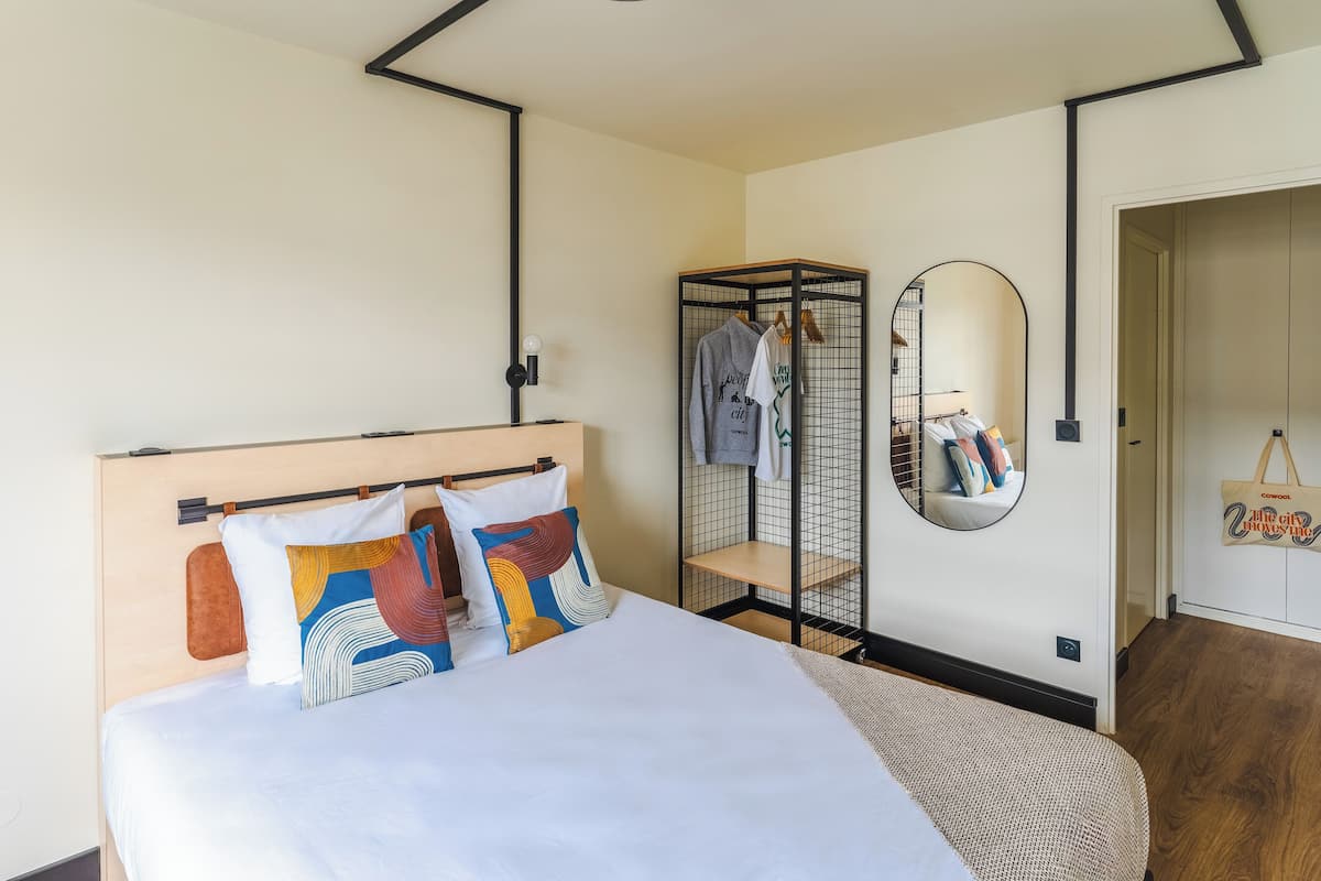 A modern hotel room with a wooden headboard, a large bed with colorful pillows, a wardrobe with hanging clothes, and a mirror on the wall.