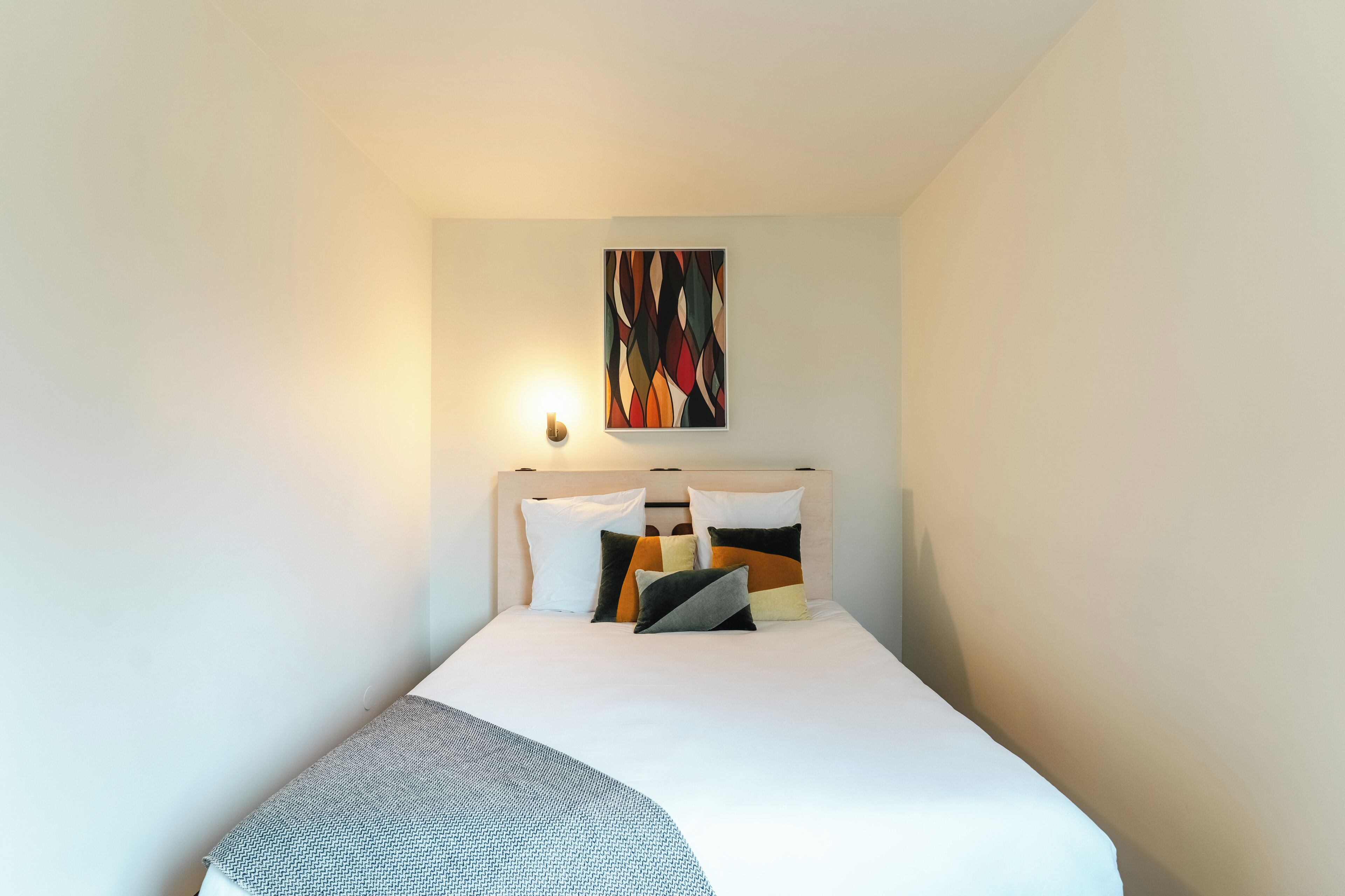 Una cama bien hecha con edredón blanco y varios almohadones, un cuadro abstracto colgado en la pared y una lámpara de noche.