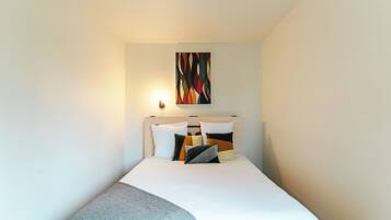 Una cama bien hecha con edredón blanco y varios almohadones, un cuadro abstracto colgado en la pared y una lámpara de noche.