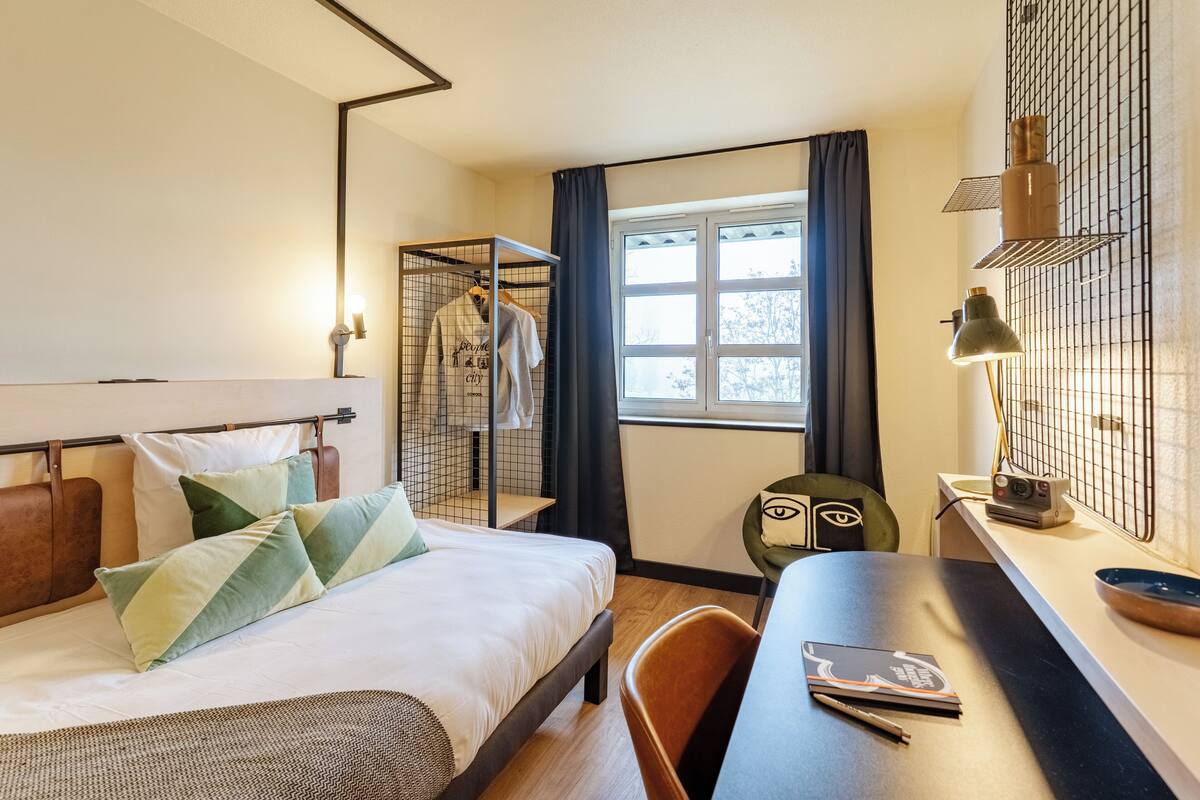 A hotel room with a bed, a desk, a chair, a window, and a wardrobe.