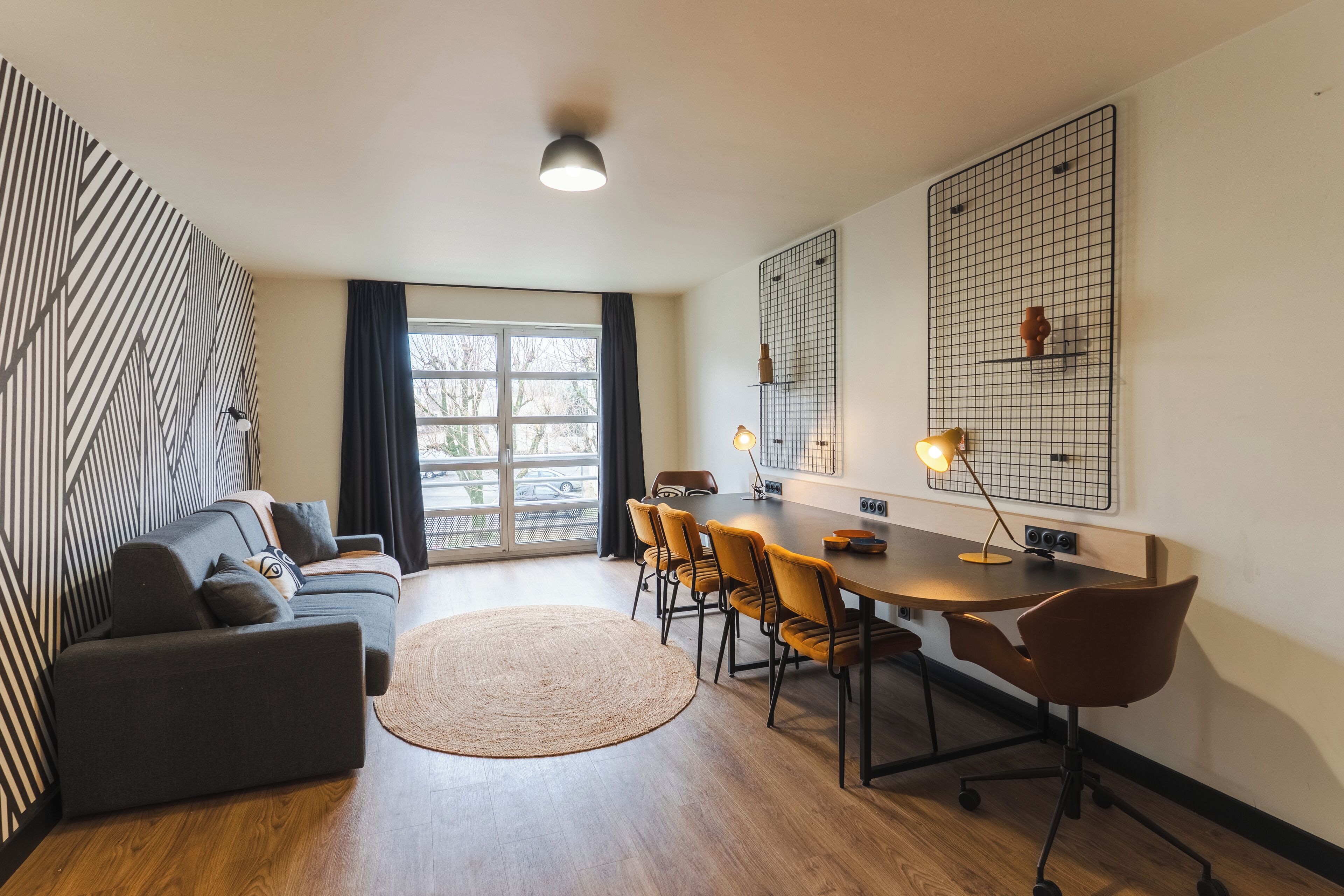 Una sala de estar moderna con un sofá, una mesa de comedor con sillas, un ventanal grande y una lámpara fijada a la pared.